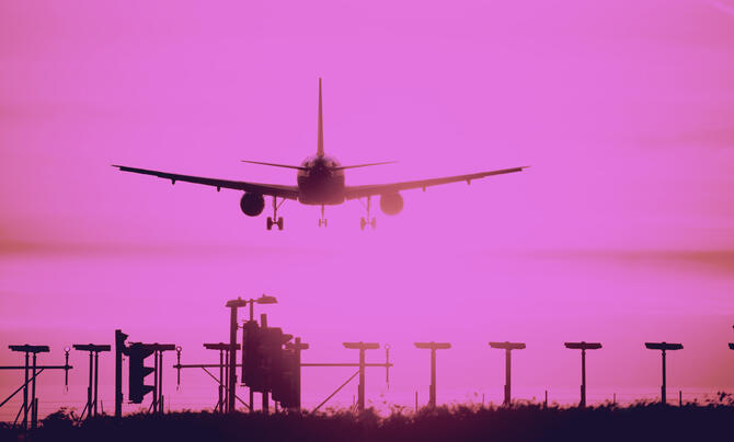 Airplane descends for landing against a vivid pink sky, silhouetted above runway lights and approach equipment.