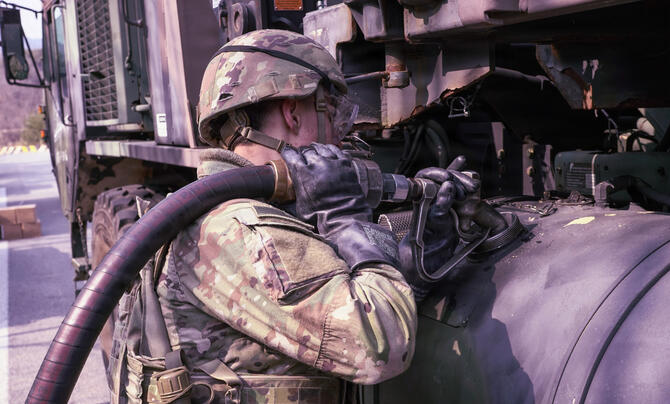 A soldier in protective gear refuels a military vehicle using a large fuel hose and heavy-duty equipment.