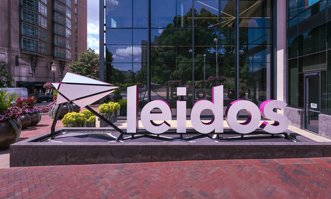 A large Leidos logo sign stands outside a modern glass office building, surrounded by landscaped greenery and brick walkways.