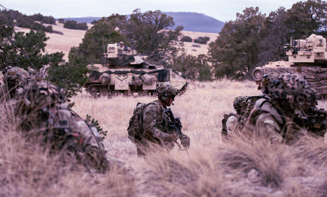 Soldiers move through rugged terrain during a military training exercise, with armored combat vehicles providing support in the background.