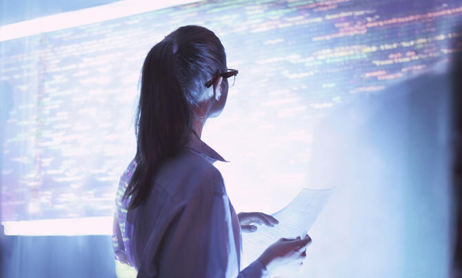 Cybersecurity professional reviews information in front of a large screen displaying computer code.