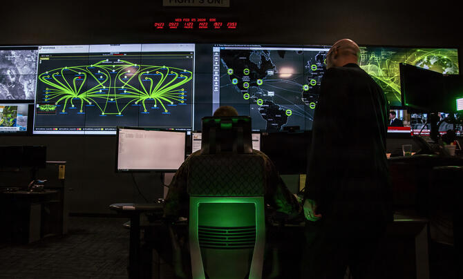 Two people facing away looking at large computer and TV screens with various data feeds in a dark room