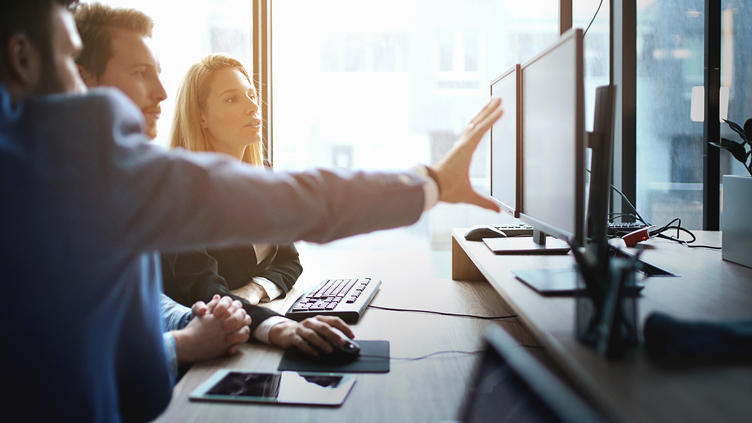 three coworkers sitting at a computer while one is pointing at a monitor